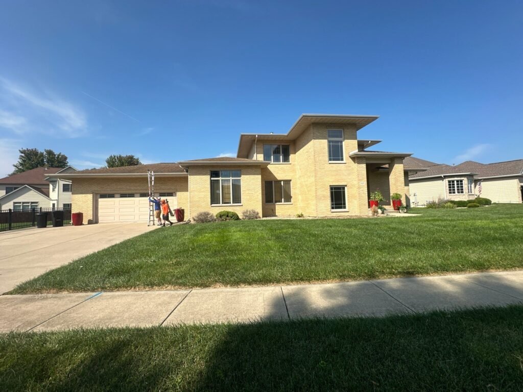 Street view of a two-story tan brick Springfield, IL home with its original brown shingle roof as the roofing crew sets up