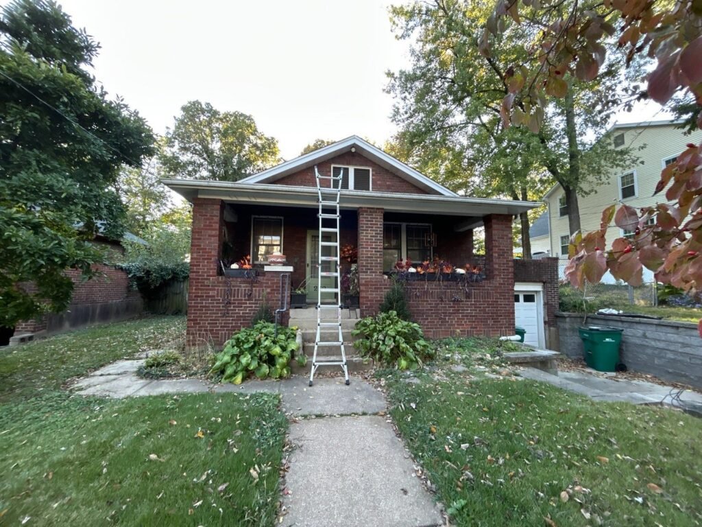 Street view of a red brick Webster Groves, MO bungalow with a ladder at the front porch before roof replacement